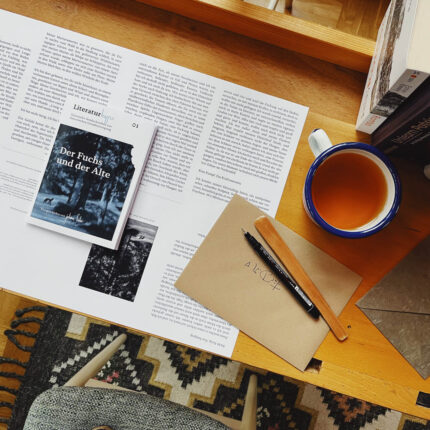 Ein Tisch mit einem Buch namens 'Der Fuchs und der Alte', einem Notizblock, einem Stift, einer Tasse Tee und gedruckten Texten. Der Tisch ist aus Holz und hat einen Teppich darunter.