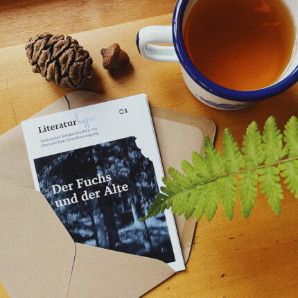 Zeitung mit dem Titel 'Der Fuchs und der Alte' auf einem Tisch, umgeben von einer Tasse Tee, einer grünen Pflanze und Dekorationselementen wie einer Tannenzapfen und einem Stein.