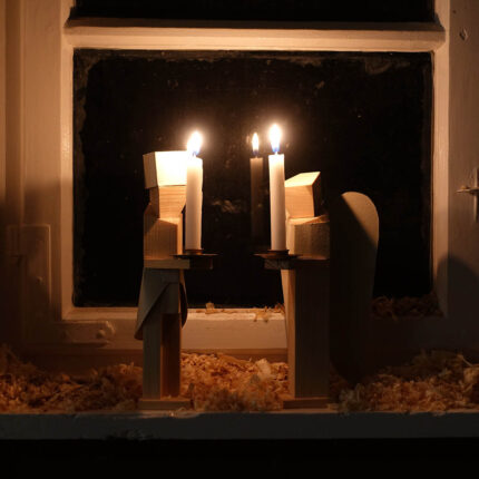 Abgebildet sind zwei kubistische Kerzenhalter aus Holz die die Form eines Engels oder Bergmanns haben. Es brennen die beiden Kerzen. Die Figuren stehen auf einem Fensterbrett, worauf Holzspähne liegen. draußen ist es dunkel.