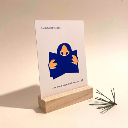 Abgebildet ist ein Schild in einem Holzaufsteller auf dem steht „Endlich mal wieder ... [blaue, stilisierte Grafik, die ein Buch zeigt, gelbe Hände und eine gelbe Nase] ... an einem neuen Buch riechen“. Daneben liegt ein kleiner Zweig eines Nadelbaumes.