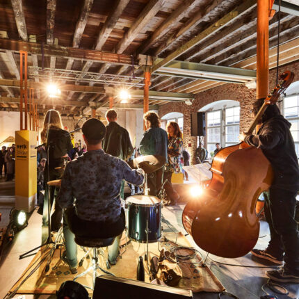 Band von hinten bei einem Auftritt in einer Industriehalle. Zu sehen ist ein Kontrabass und ein Schlagzeug mit Instrumentalist:in von hinten.